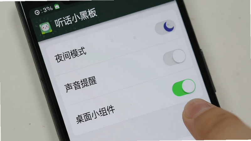 展示一部安卓手机屏幕,显示“听话小黑板”APP的设置页面。页面中有几个开关选项,如“夜间模式”、“声音提醒”、“桌面小组件”。手指正点击“桌面小组件”开关,开关变为绿色开启状态。背景为浅灰色,界面清晰,字体易读,风格为写实UI截图,高清,无杂乱背景。