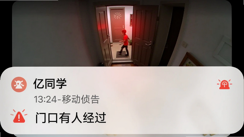 主体为手机通知栏弹出“小亿同学”移动侦测告警消息;布局上半部分为告警画面缩略图,下半部分文字提示“门口有人经过”;风格简洁通知界面;细节包括时间戳、设备名称、红色告警图标;画质清晰干净。