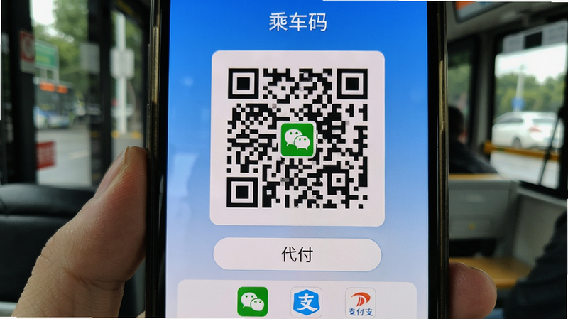 月城公交扫码支付界面,手机屏幕显示一个大型二维码,上方有“乘车码”标题,下方有“代付”按钮和支付方式图标(微信、支付宝),背景为蓝色渐变,简洁干净