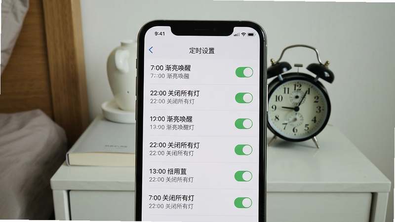 手机屏幕显示定时设置页面,列表中有多条定时任务(如“7:00 渐亮唤醒”、“22:00 关闭所有灯”),背景是卧室床头柜,上面有一个闹钟。构图居中,风格简洁,细节包括定时表格的线条和开关图标,画质干净,无多余元素。