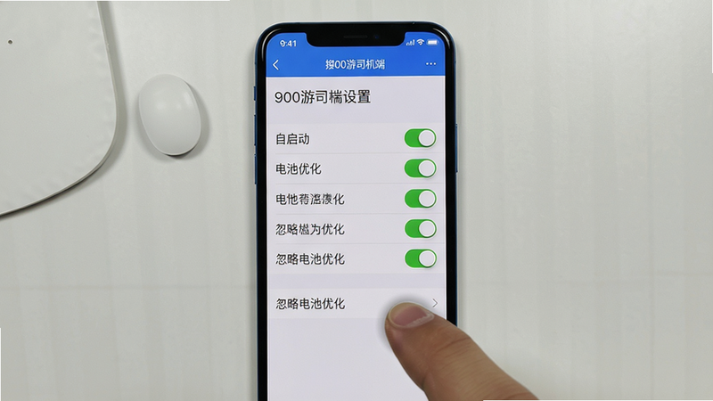主体:手机竖屏显示900游司机端设置页面,有“自启动”、“电池优化”等选项开关。布局:手机居中放置,桌面背景为灰白色。排版:开关按钮为绿色开启状态,选项文字清晰。风格:UI截图风格,简洁真实。细节:手指虚影正在点击“忽略电池优化”。画质:高清,无多余元素。