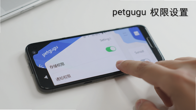 主体为一部安卓手机平放在桌面上,屏幕显示 petgugu 应用的设置页面,其中“存储权限”和“通知权限”开关处于开启状态,旁边有一只手正在点击,背景干净,风格写实,画质高,文字标注“petgugu 权限设置”。