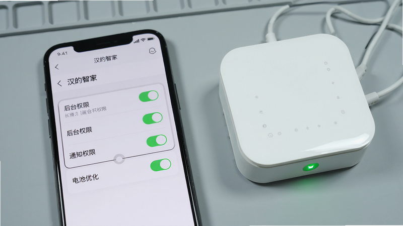 手机屏幕显示“汉的智家” App 内设置页面,突出显示“后台权限”“通知权限”“电池优化”三个选项,每个选项旁边有开关按钮,其中两个已打开。手机旁边放置了一个方形智能网关,指示灯为绿色。背景为浅灰色工作台,画质细腻,无杂乱元素