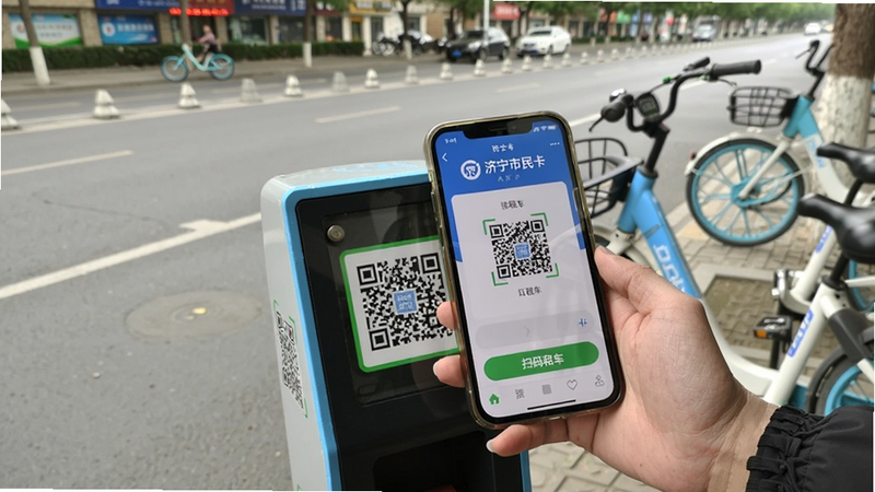 һ����Ƭ��ʾ�û��ֳ��ֻ�ɨ�蹫�����г�׮λ�ϵĶ�ά���ֻ���Ļ����ʾ��������APP���⳵���汳��Ϊ�ֵ������г�����Ϊ�ֻ���׮λ���־����Ű���ʵ����������ϸ�ڰ����ֻ���Ļ�ϵġ�ɨ���⳵����ť����������Ȼ
