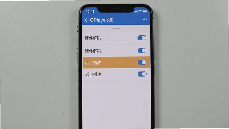 画面主体:一部手机竖屏显示OPlayer设置菜单,突出显示“硬件解码”、“后台播放”等选项。布局:手机居中,背景为浅灰色。风格:真实截图风格,顶部有状态栏。细节:每个选项旁边有开关按钮,界面中文。画质:清晰,无多余元素。