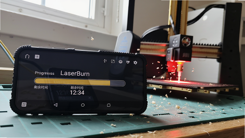 主体:一台安卓手机横屏放置在工作台上,屏幕显示LaserBurn正在雕刻进度条,旁边有激光雕刻机正在工作,发出红光。布局:手机在左前,雕刻机在右后,视角斜45度。排版:手机屏幕上可见“剩余时间 12:34”字样。风格:实拍风格,自然光线。细节:木屑飞扬,手机壳有防滑纹路。画质:1080p,干净,无多余元素。