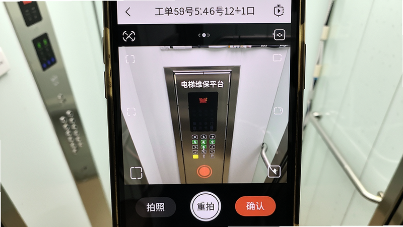 一部手机拍摄电梯控制柜的俯视特写,手机屏幕显示“电梯维保平台”拍照界面,取景框内有电梯控制柜局部,屏幕底部有“拍照”“重拍”“确认”按钮,顶部显示工单编号。背景为电梯机房,光线充足,画面清晰,干净整洁。