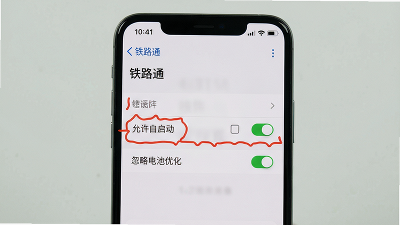主体:手机屏幕显示“铁路通”设置界面,高亮显示“允许自启动”和“忽略电池优化”两个选项。布局:手机居中,界面为系统设置页,选项前有红色标注。排版:文字清晰,开关按钮为绿色开启状态。风格:手机系统截图风格,真实感。细节:状态栏显示时间,背景为浅色。画质:高分辨率,无多余元素。