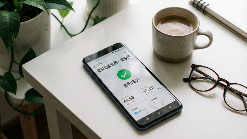 主体为一部安卓手机平放在桌面上,屏幕显示“聊天记录免费一键备份”的备份完成界面,界面打上绿色的对勾并显示“备份成功”字样。手机旁边摆放着一杯咖啡和一副眼镜,营造出轻松日常的使用氛围。背景是简约的书桌与绿植,布局采用俯拍视角,排版自然随意,风格为生活化摄影写实,细节上突出屏幕内的数据统计数字,画质柔和,景深浅,干净无多余元素。