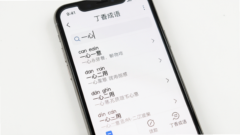 手机屏幕显示“丁香成语”搜索界面,搜索框内有“一心”二字,下方列表展示“一心一意”“一心二用”等成语,每条带有拼音和简释。背景为白色,底部有功能图标,整体布局简洁清晰,风格干净明亮,画质高清,无多余元素。