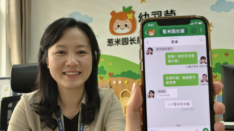 主体:一位园长正在使用手机与家长进行视频通话,手机屏幕上同时显示着葱米园长版的聊天界面。布局:人物位于画面左侧三分之一处,手机屏幕朝向镜头。排版:前景是手机屏幕特写,背景是园长的办公场景。风格:写实生活照风格,自然光,画面温馨。细节:手机屏幕上可见对话气泡和“葱米园长版”字样,园长表情和蔼,背景有幼儿园的LOGO墙。画质:高清,人脸清晰,干净无多余元素。