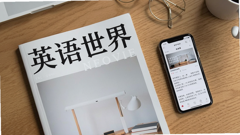 主体:一本《英语世界》杂志平铺在桌面上,旁边放着一部手机,手机屏幕显示应用内文章;布局:杂志与手机并排,背景为书桌;排版:俯拍视角;风格:简约文艺;细节:杂志封面有“英语世界”四个字,手机屏幕有应用界面;画质:高清,干净无多余元素