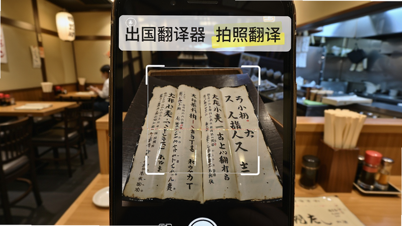 主体为手机相机取景界面,布局中心为一张日式菜单照片,上方叠加半透明白色翻译文本框,背景为模糊餐厅环境,风格写实摄影风,细节展示对焦框与翻译高亮效果,文字包含