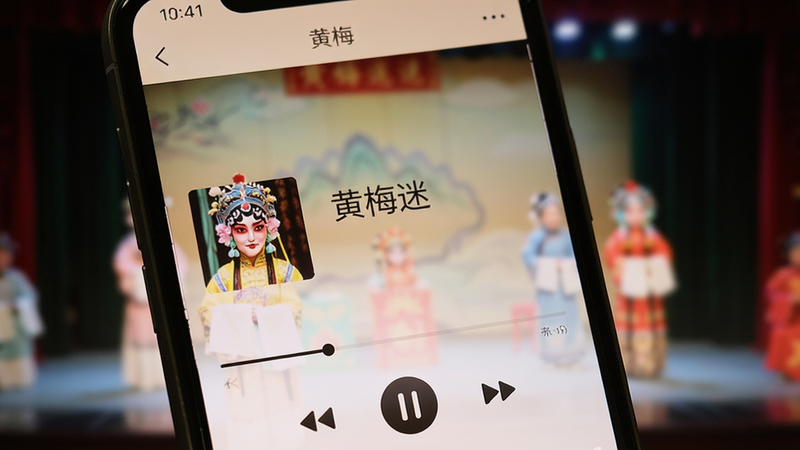 手机屏幕特写,显示“黄梅迷”APP的播放界面,背景为模糊的戏曲舞台,中间显示当前播放曲目名称,下方有进度条和控制按钮,画面清晰,色彩柔和,体现沉浸式体验