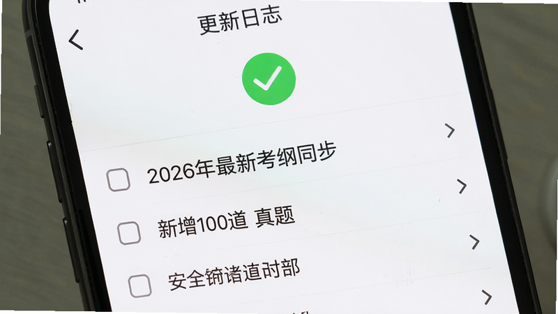 手机屏幕显示“更新日志”页面,顶部有绿色对勾图标,下方列出“2026年最新考纲同步”、“新增100道真题”等条目,背景为白色,字体深灰,排版整齐,高清细节