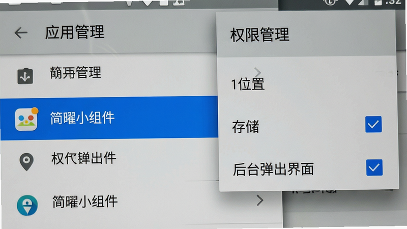 手机设置界面模拟图,展示“应用管理”列表,其中“简曜小组件”条目被高亮选中。右侧弹出“权限管理”子菜单,显示“位置”、“存储”、“后台弹出界面”三个选项,其中“后台弹出界面”勾选状态清晰可见。界面采用标准安卓Material Design风格,灰色背景,蓝色选中态,文字清晰无遮挡。风格写实,模拟真实手机屏幕拍摄,画质高,无摩尔纹,干净无多余图标。